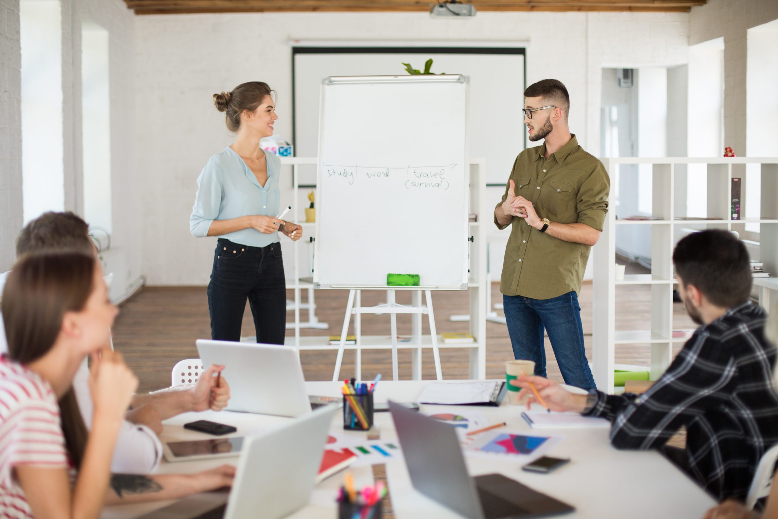 Man in eyeglasses and shirt and smiling woman in blouse standing near board dreamily presenting new project to colleagues. Group of young people working together in modern white office